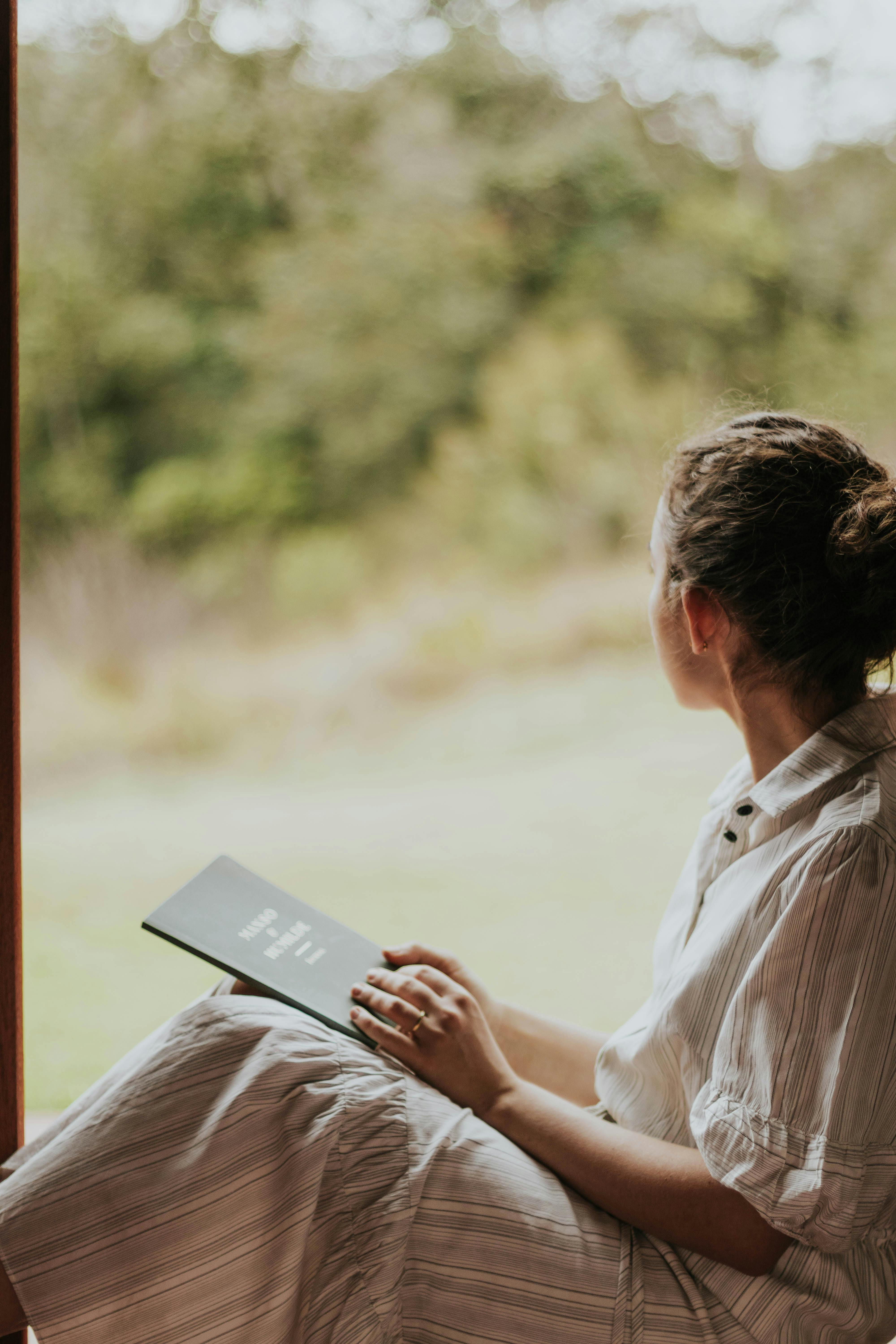 A woman in a white dress enjoys reading a book by a window overlooking a serene countryside landscape.