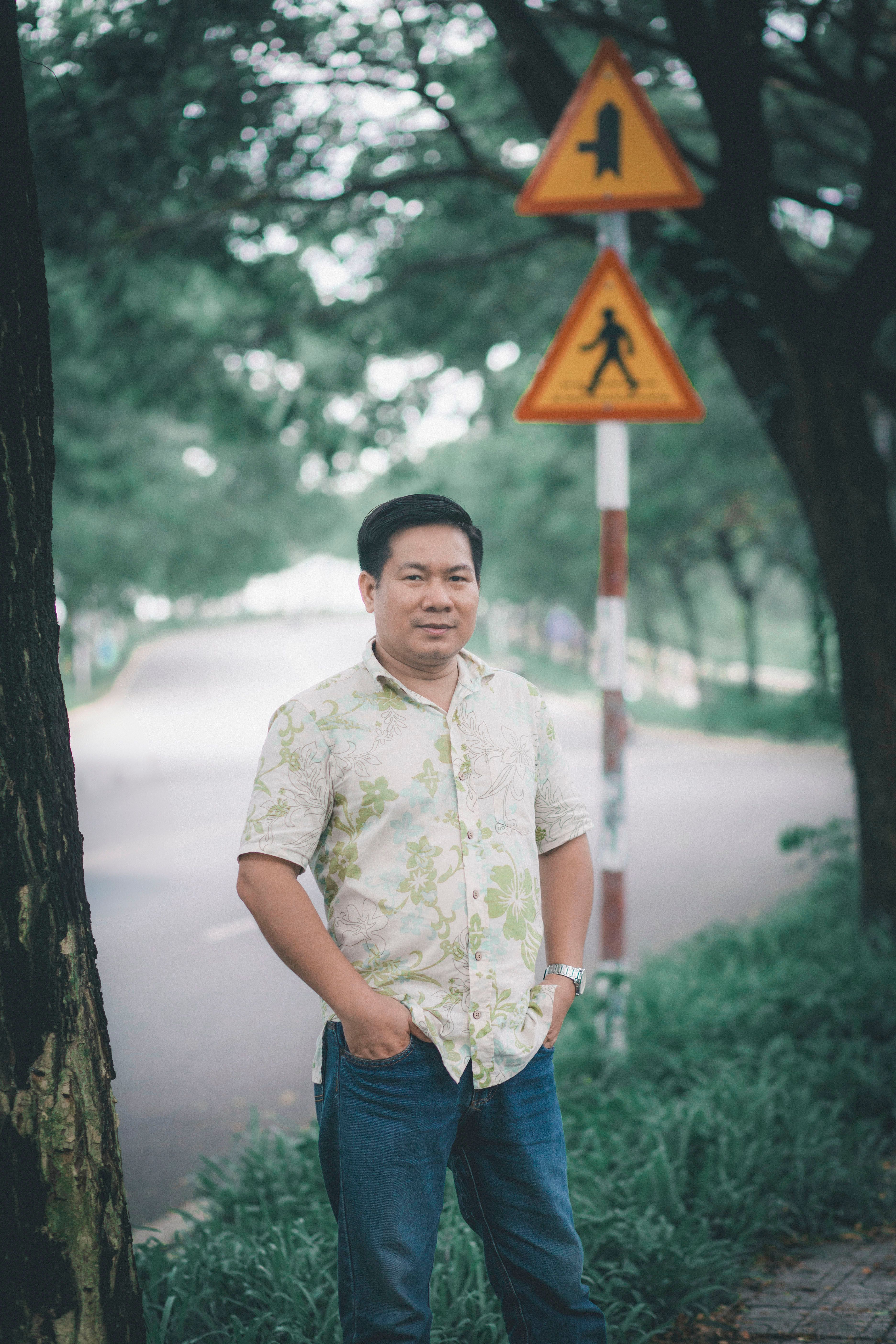 Asian man in floral shirt standing casually outdoors with trees and traffic signs in the background.