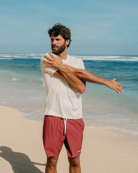 Adult man in sportswear stretching by the sea on a sunny day, dressed casually in a t-shirt and shorts.