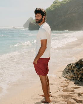 Casual man in white shirt and red shorts standing on a sunny beach by the ocean.