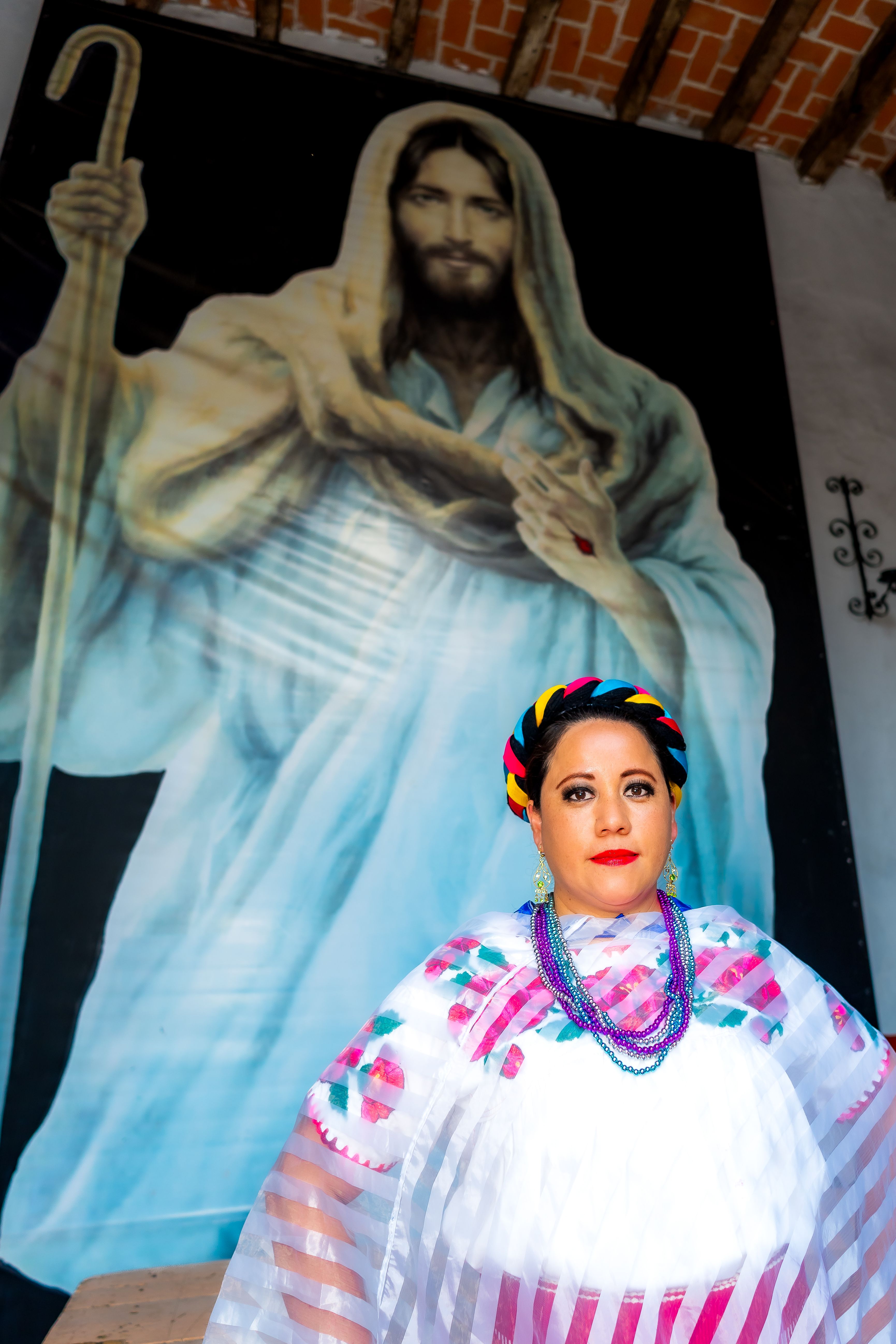 Woman in traditional Mexican attire poses with a religious mural background.