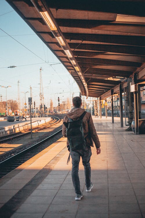 Vista Traseira Do Homem Fotográfico Com Capuz Marrom, Calça Cinza E Mochila Andando Sozinho Na Plataforma Vazia Da Estação Ferroviária