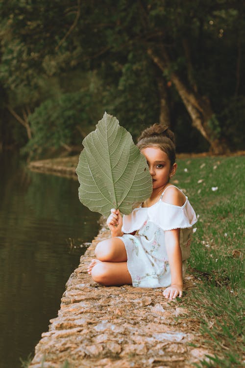 Free Girl Holding Green Leaf Sitting Near Body of Water Stock Photo