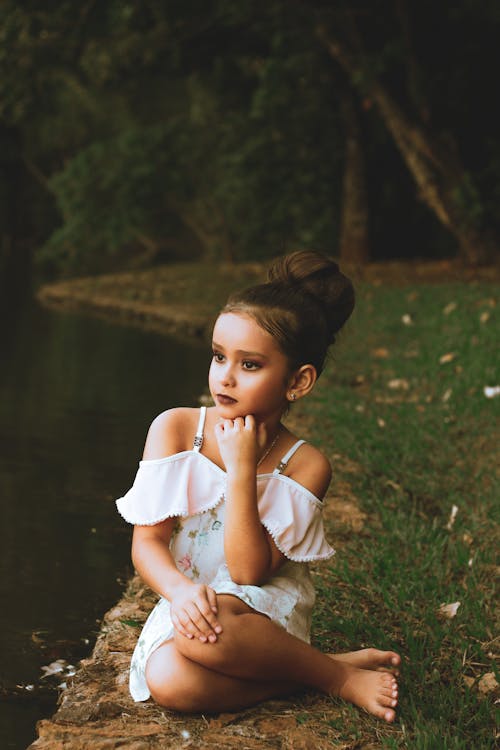 Free Photo of Young Girl in Make-up Wearing White Off-shoulder Dress Sitting on Grass Near Body of Water Looking Away Stock Photo