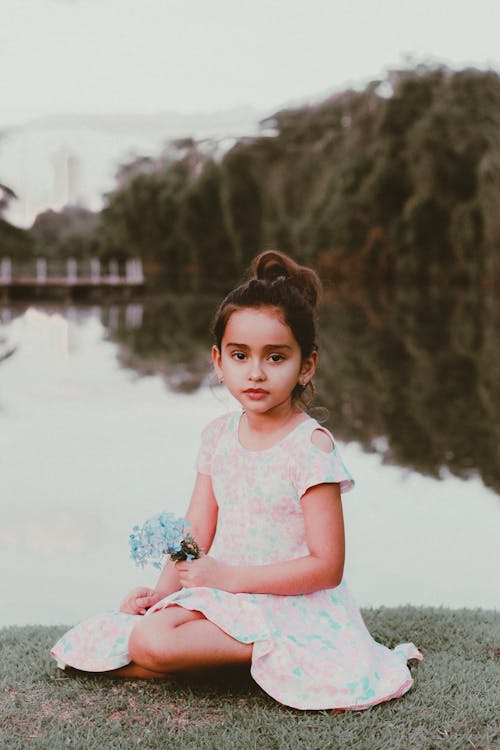 年轻的女孩穿着白色花裙子的化妆，坐在水边的草地上，同时手捧鲜花的照片