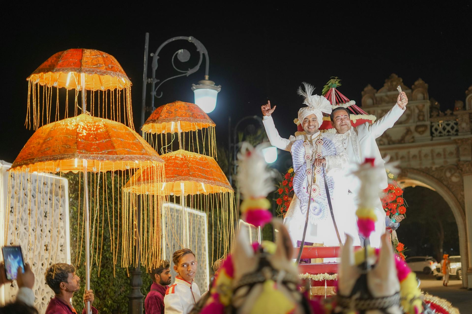 A vibrant Indian wedding procession featuring the groom in traditional attire, celebrating under colorful parasols at night.