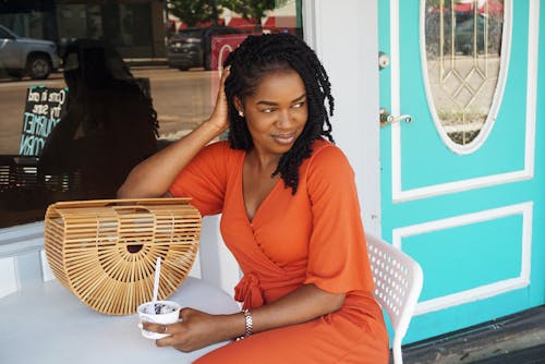Photo of Smiling Woman in Orange V-neck Short-sleeved Dress Sitting Outside Store Holding Ice Cream While Looking Away 