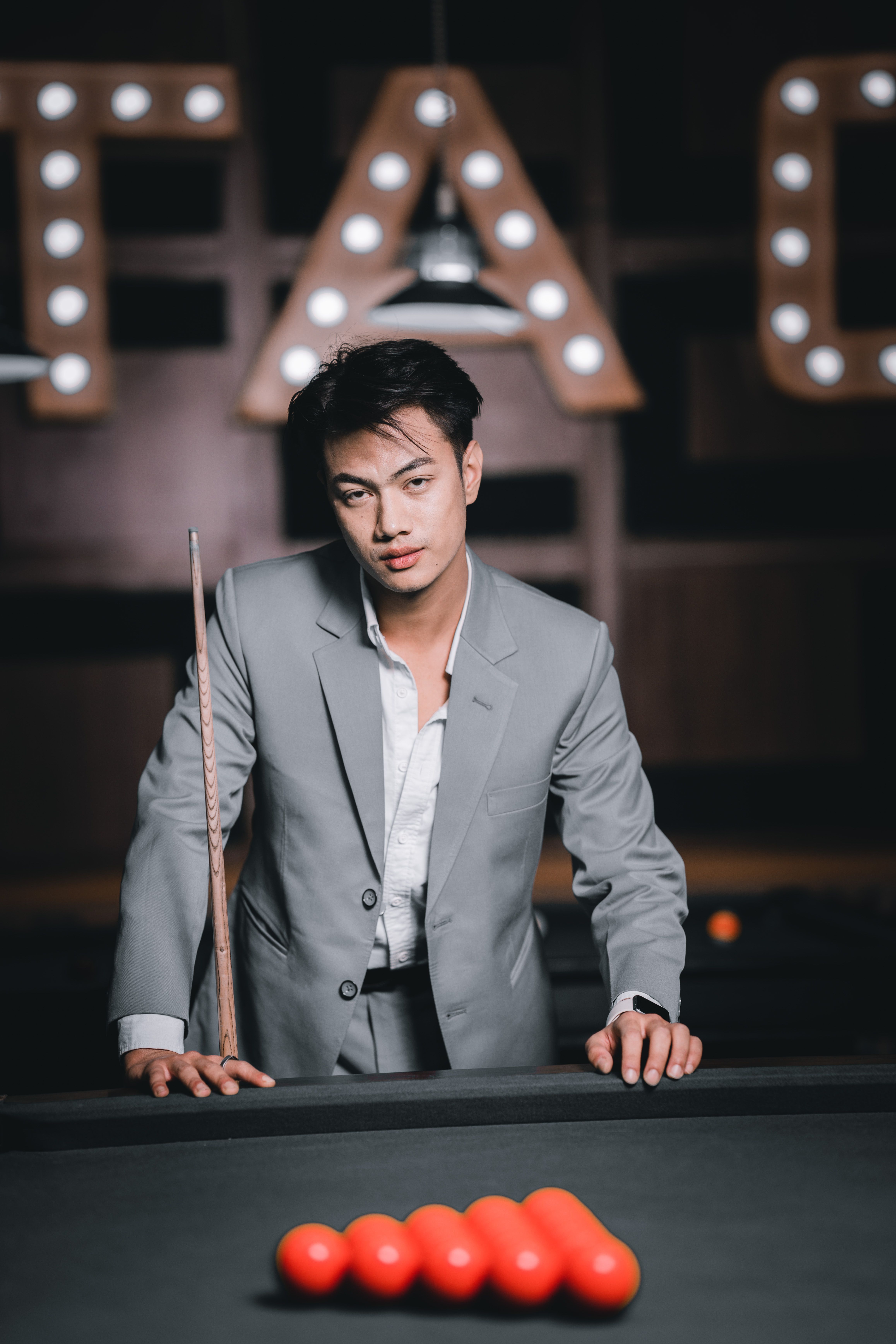 Stylish man in a grey suit holding a cue stick, posing by a pool table with red balls.