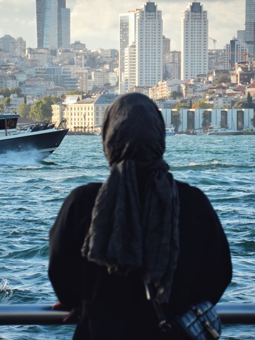 Free Woman in hijab standing by the Bosphorus Strait, observing the modern Istanbul skyline with skyscrapers. Stock Photo
