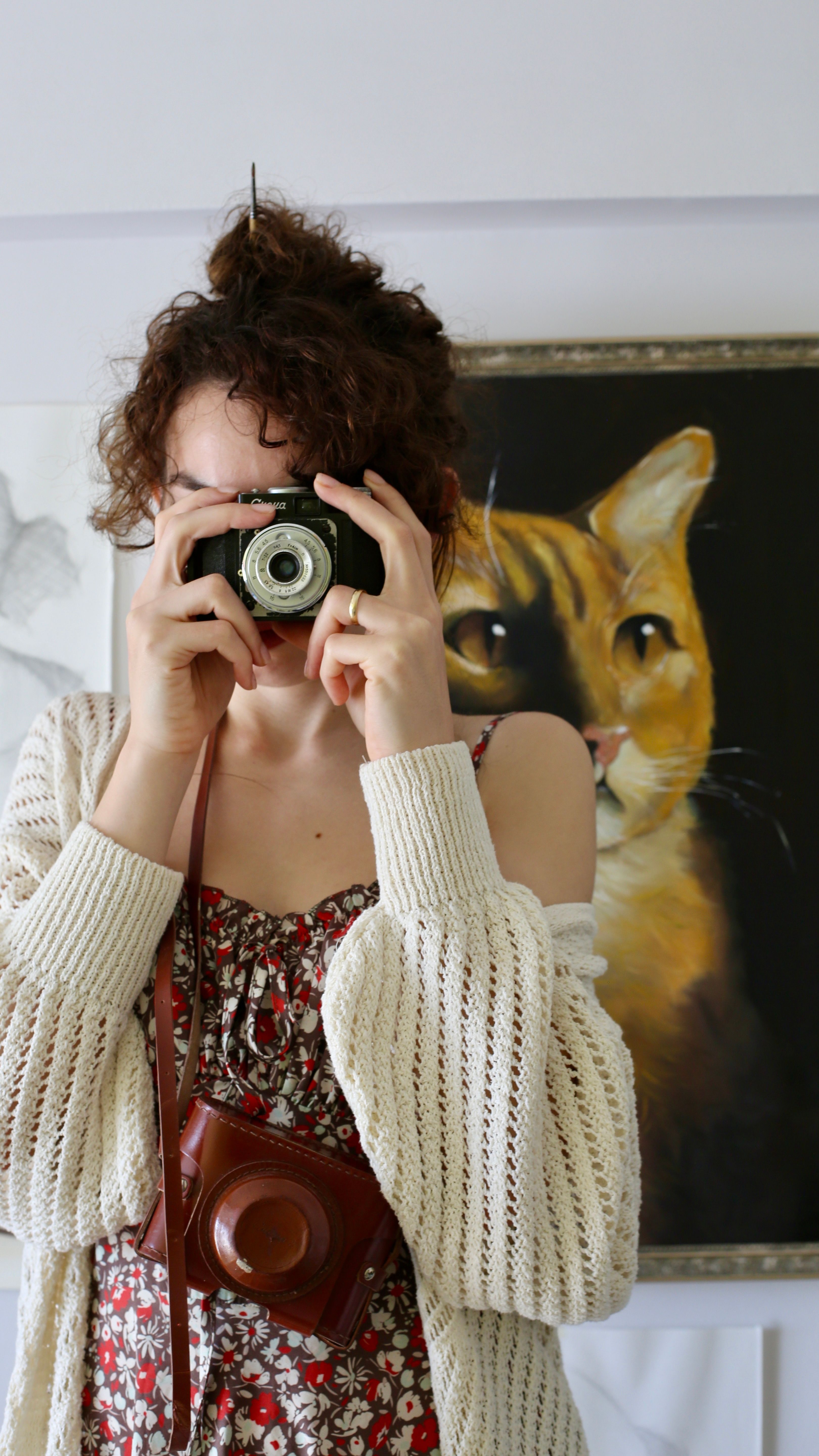 Young woman in floral dress and cardigan taking a photo indoors with a vintage camera.