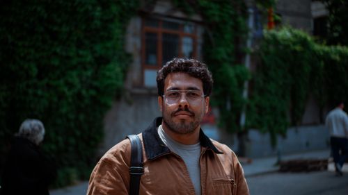 Free A man stands on a city sidewalk wearing eyeglasses, exuding a confident urban vibe. Stock Photo