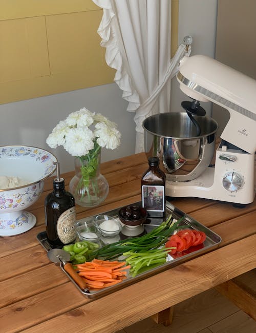 A kitchen table with a mixer, vegetables and a bowl of pasta