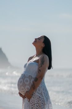 Pregnant woman in a white lace dress enjoying a serene moment by the beach.