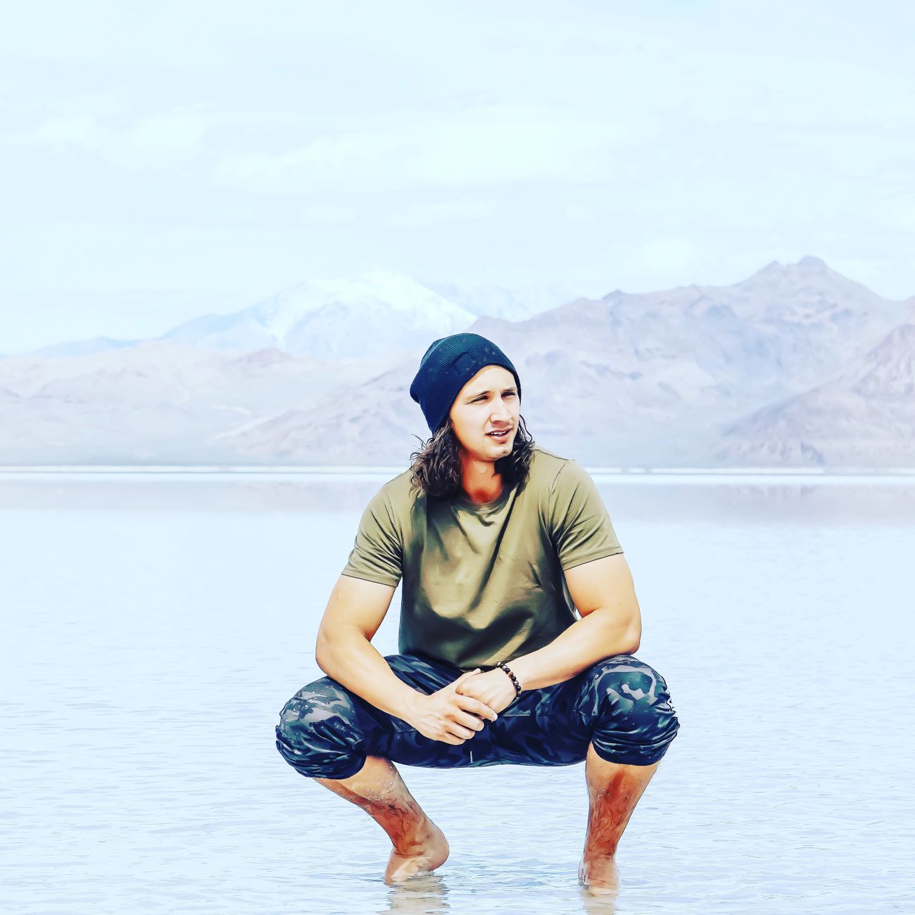 Young man with long hair in beanie squatting by a mountain lake, enjoying leisure in nature.