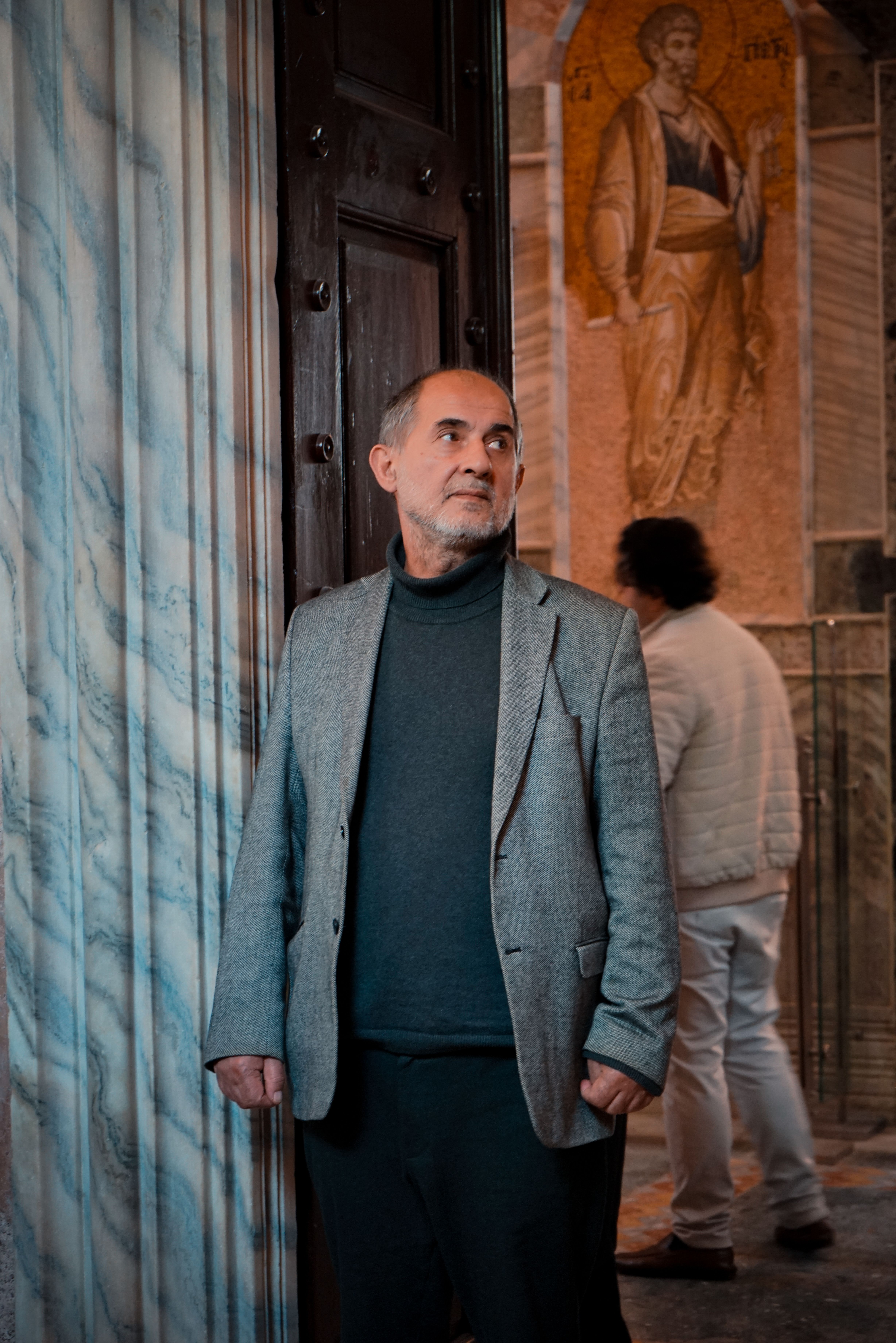Senior man in gray suit jacket stands inside a historic, ornate building with marble walls.