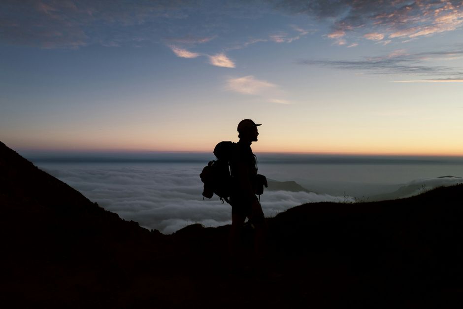 Silhouette of a hiker on a mountain trail at sunset with a sea of clouds below.