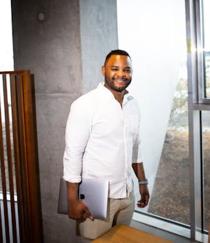 Smiling man in white shirt holding laptop stands by window in office setting.