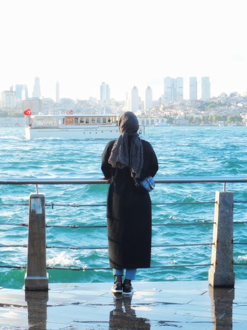 Free A woman in a hijab stands by the Bosphorus Strait, gazing at Istanbul's skyline. Stock Photo