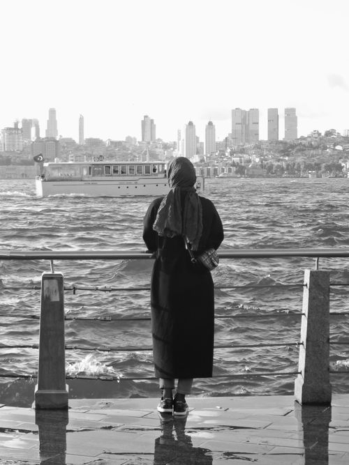 Free A woman in a headscarf standing on a promenade, gazing at the city skyline across the sea. Stock Photo