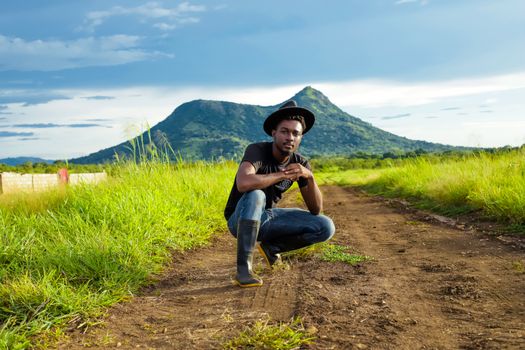 A young man squatting on a dirt road with a mountain and vibrant greenery in the background.