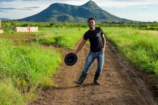 Young man holding a hat, posing on a dirt path in vibrant African countryside.