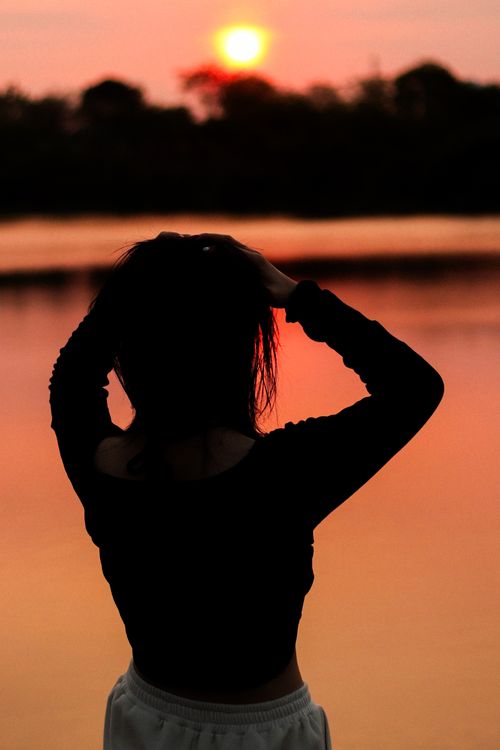 Free A silhouette of a woman stands by a serene lake during sunset, capturing the peaceful reflection. Stock Photo