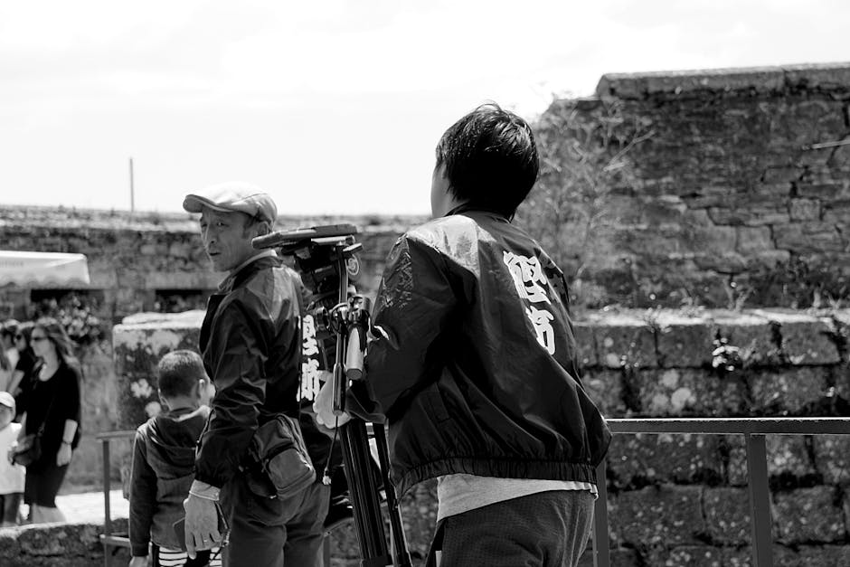Black and white image of a cameraman filming near stone walls outdoors, with people in the background.
