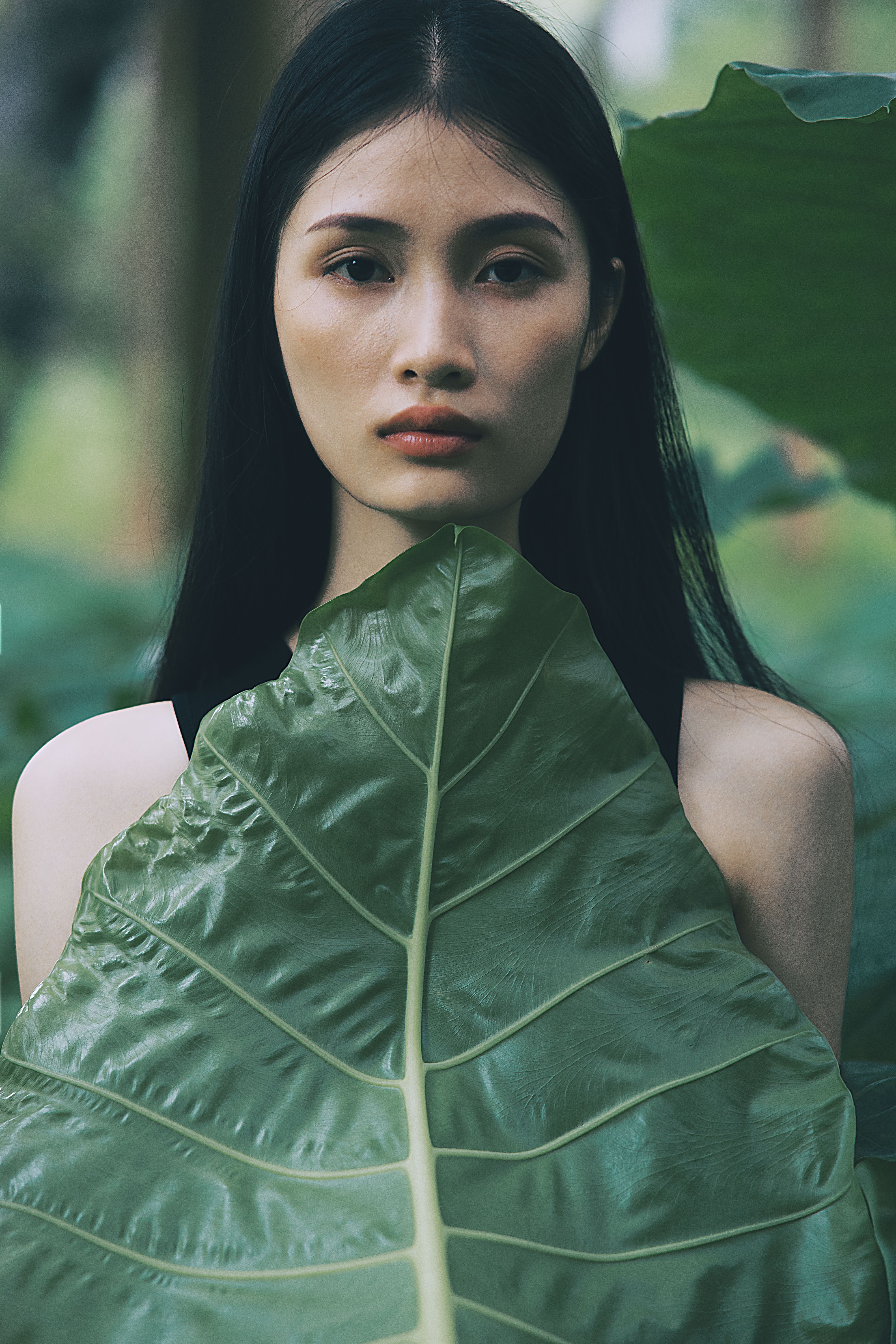 Portrait of a young woman with dark hair behind a large green tropical leaf, evoking calm and nature.