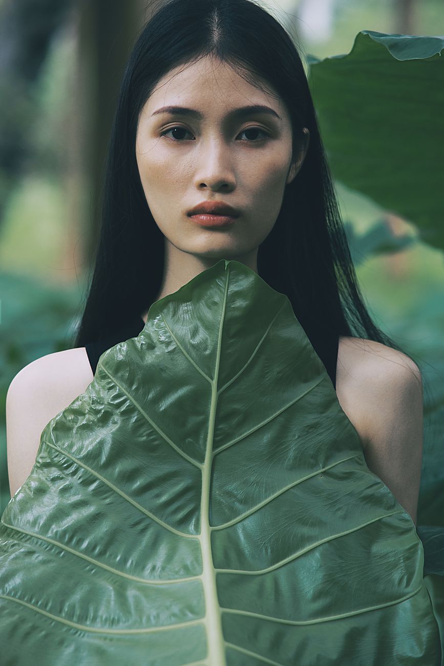 Portrait of a young woman with dark hair behind a large green tropical leaf, evoking calm and nature.