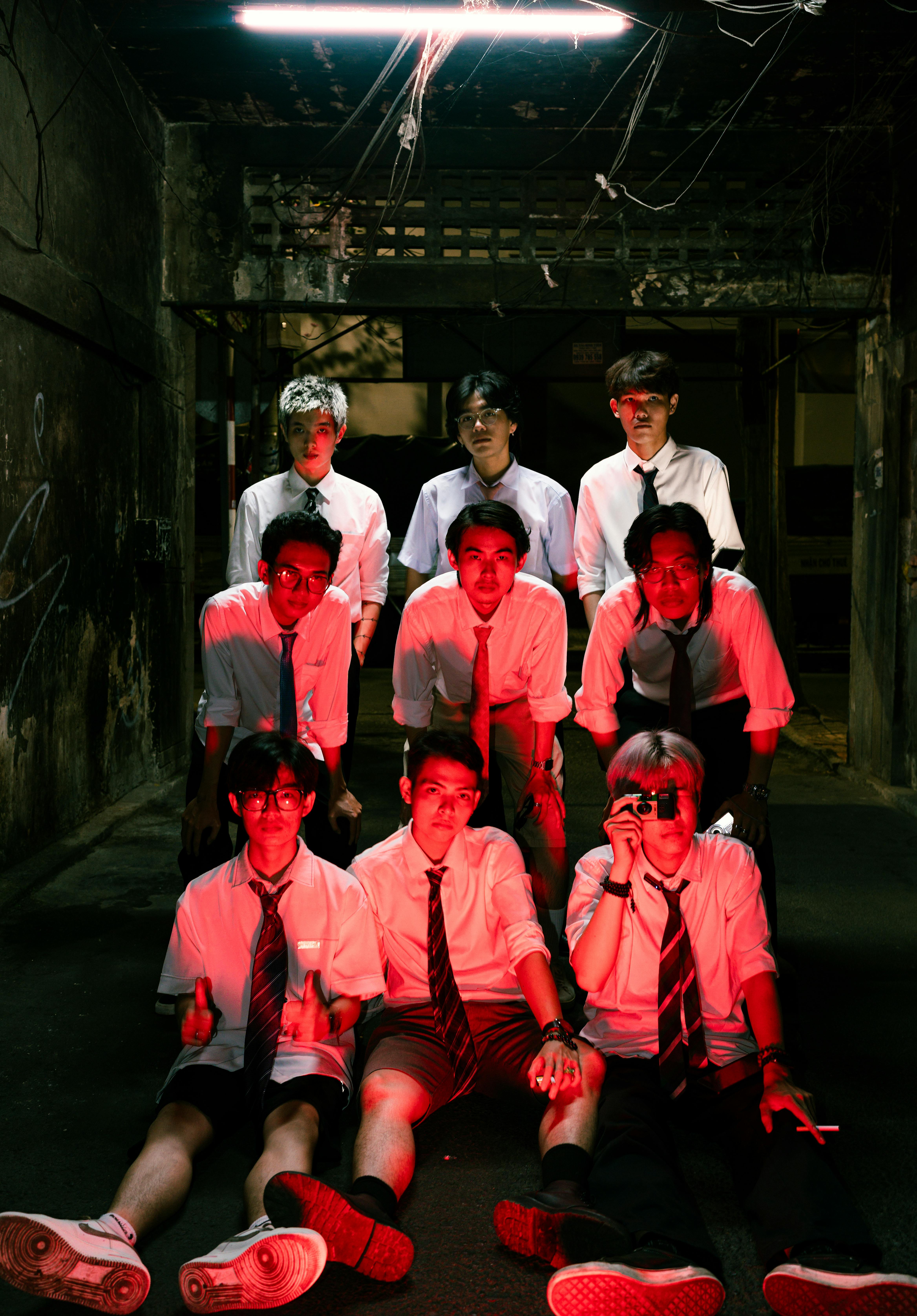 Nine men in shirts and ties under dramatic red lighting in an urban alleyway.
