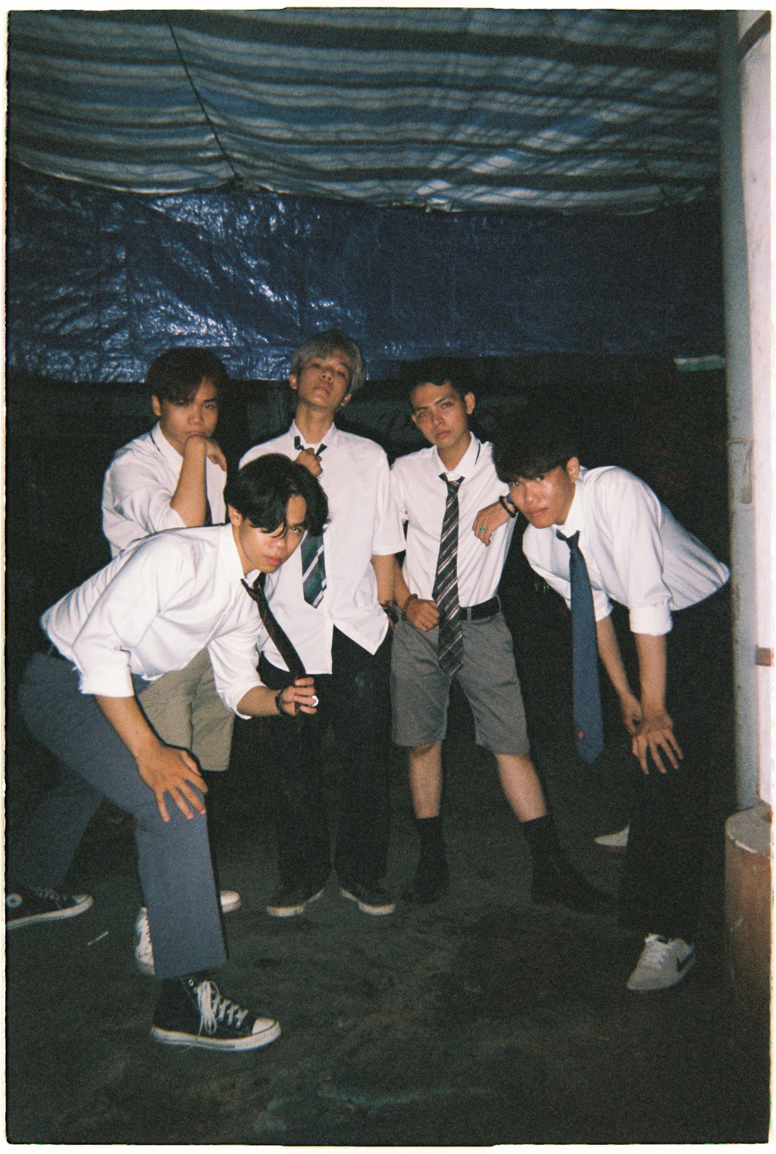 Five young men posing casually in a dark indoor setting wearing white shirts and ties.