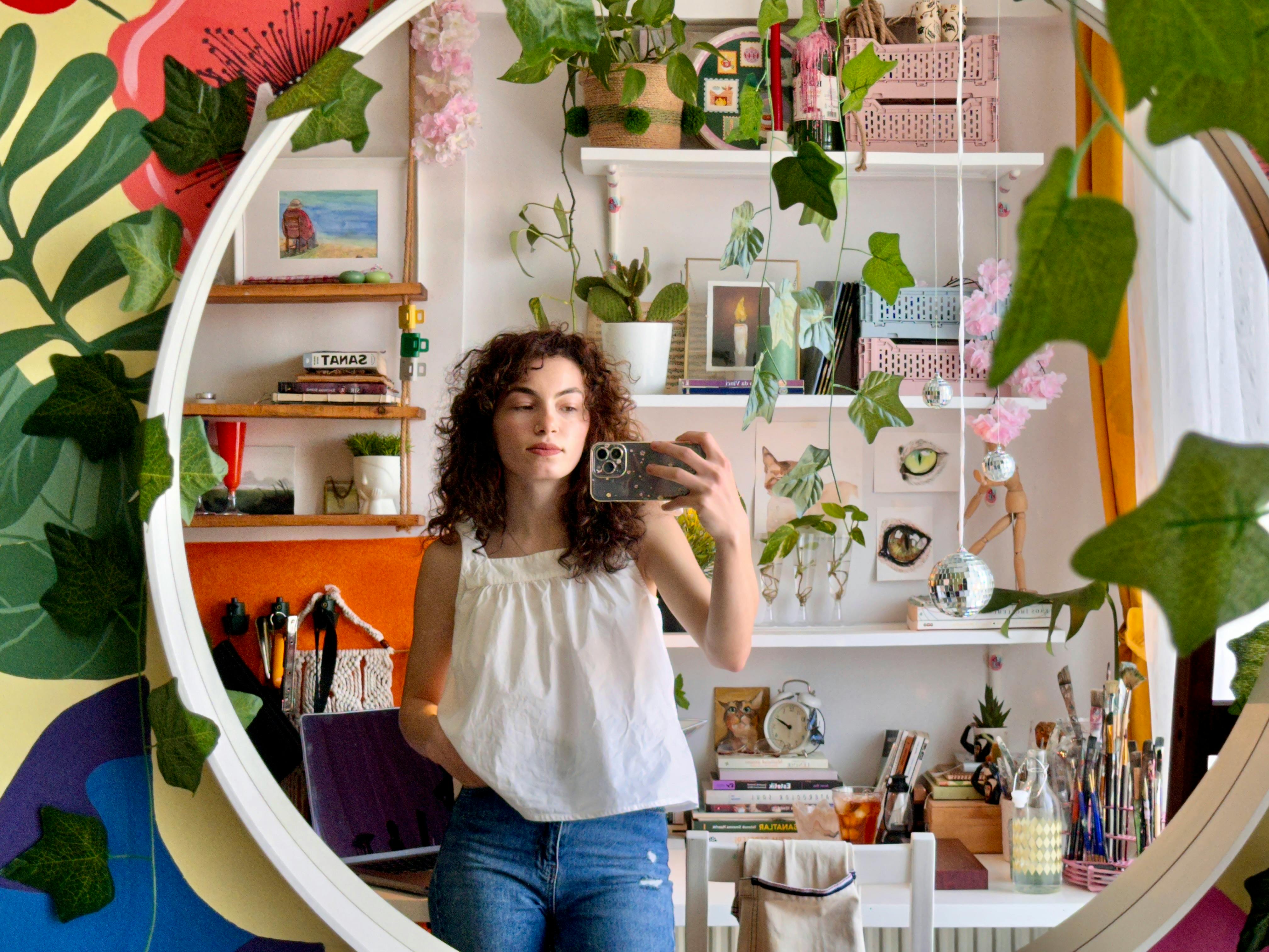 Woman taking a selfie in an art studio mirror surrounded by plants and art supplies.