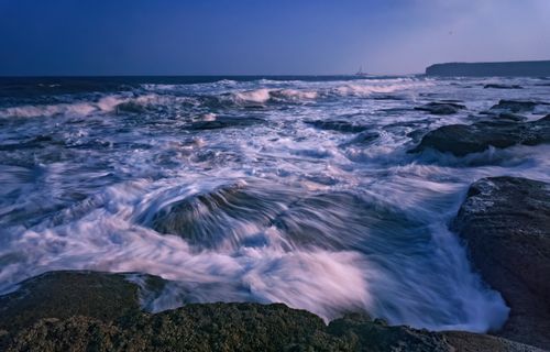 Free A mesmerizing scene of waves crashing over rocks during twilight at the seashore. Stock Photo