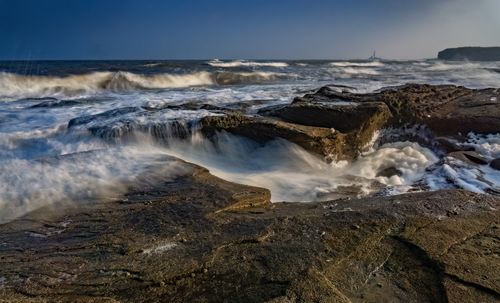Free A stunning view of waves crashing over rocks at Whitley Bay with a distant lighthouse. Stock Photo