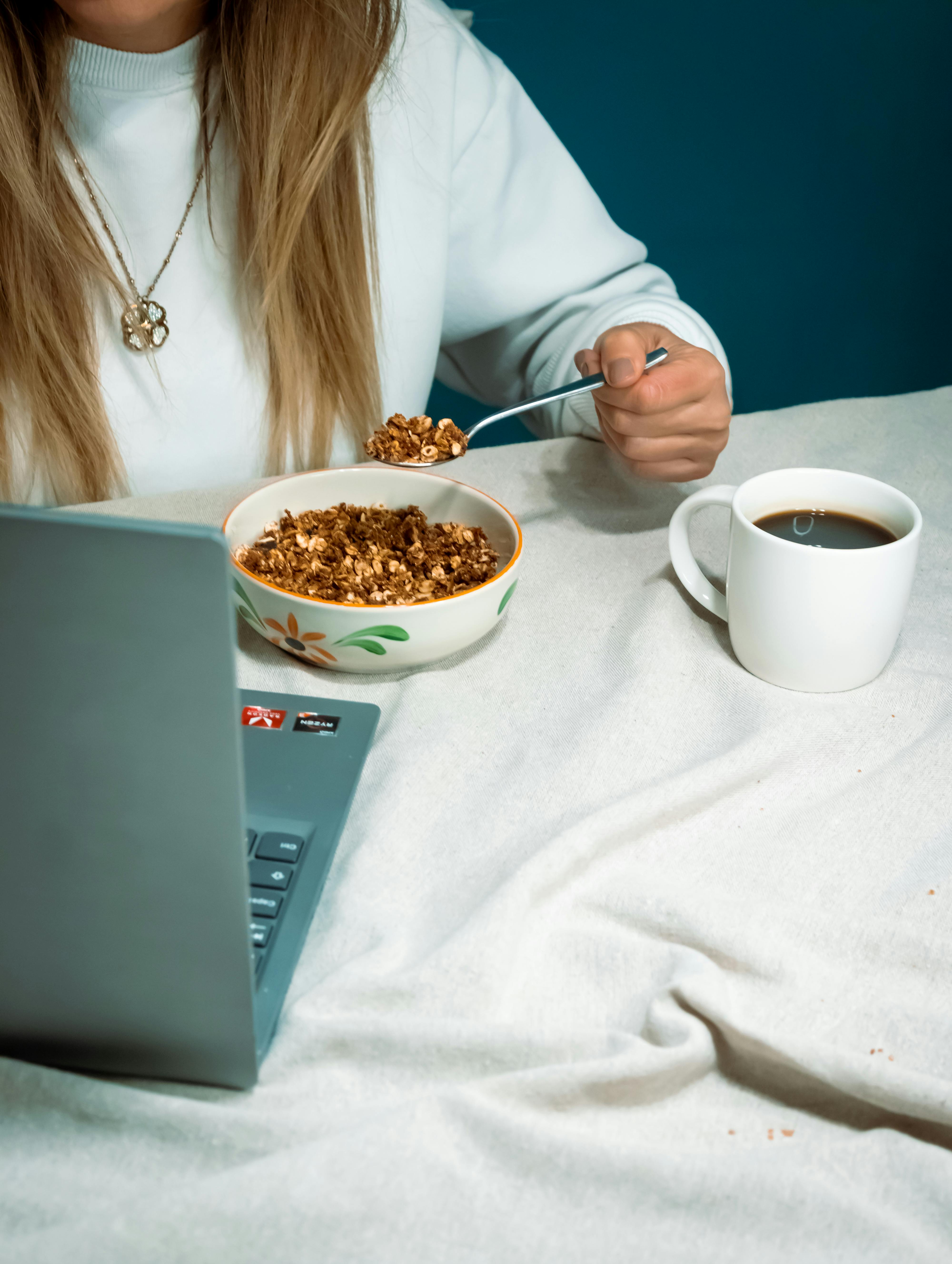 Faceless woman eats breakfast while working on a laptop with coffee, indoors.