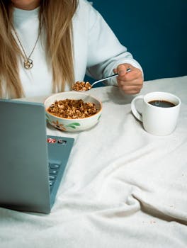 Faceless woman eats breakfast while working on a laptop with coffee, indoors.