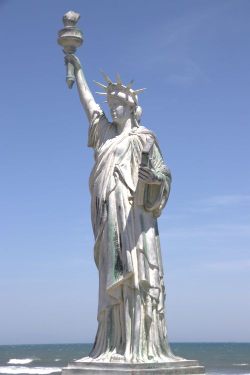 Free Replica of the Statue of Liberty against a bright blue sky and ocean background. Stock Photo