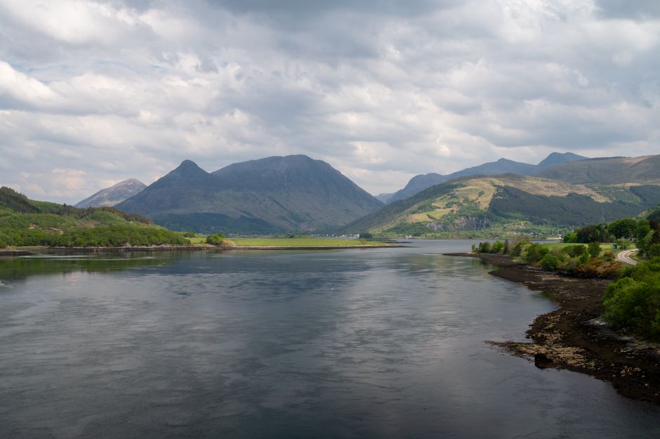 A serene landscape of Loch Leven surrounded by mountains under a cloudy sky in the Scottish Highlands.