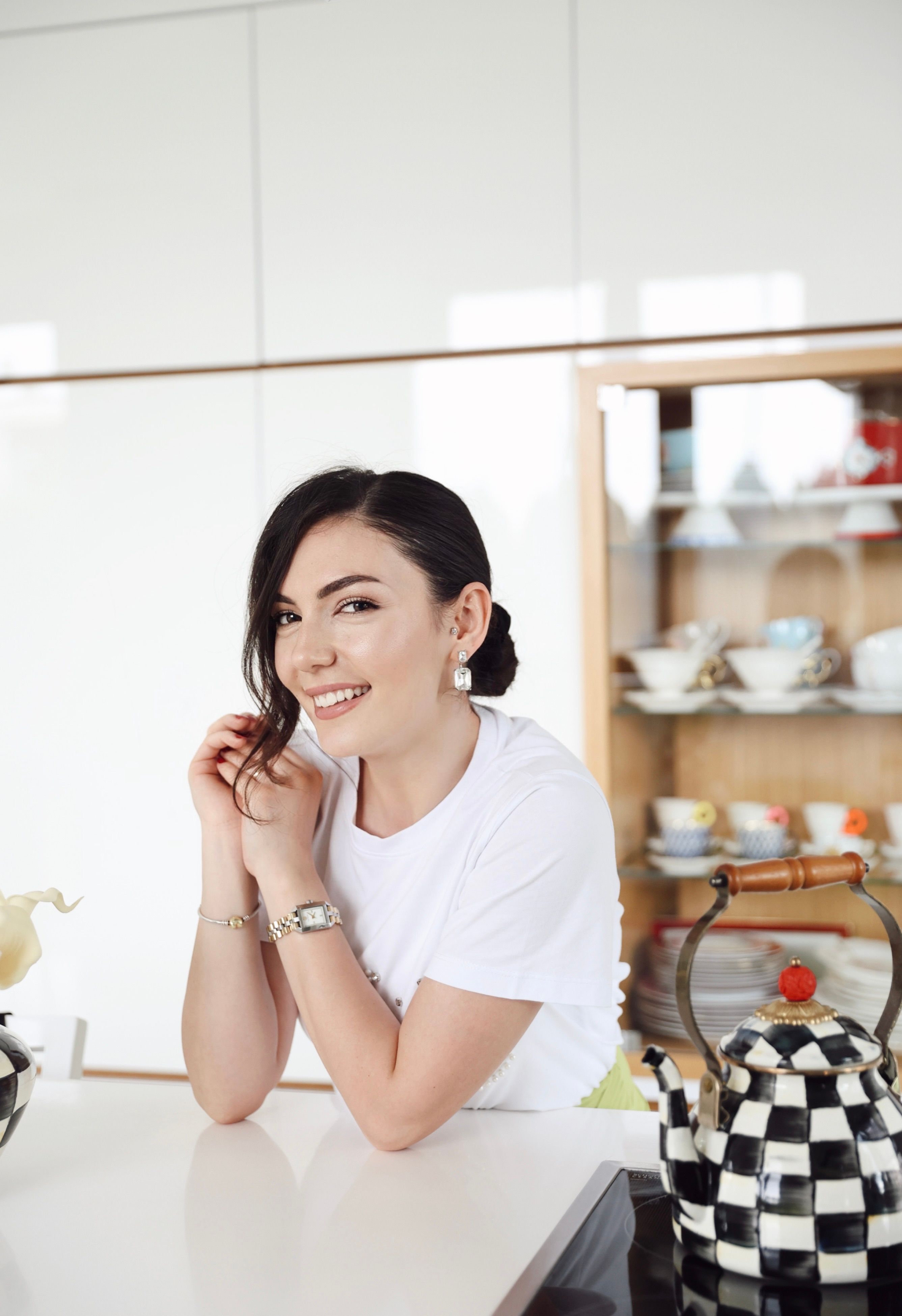 A smiling woman in a white t-shirt leans on a kitchen counter beside stylish kettles.