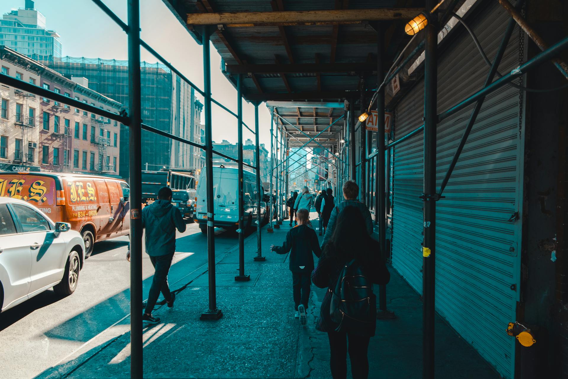 City street scene showing people walking under construction scaffolding with vehicles on a sunny day.