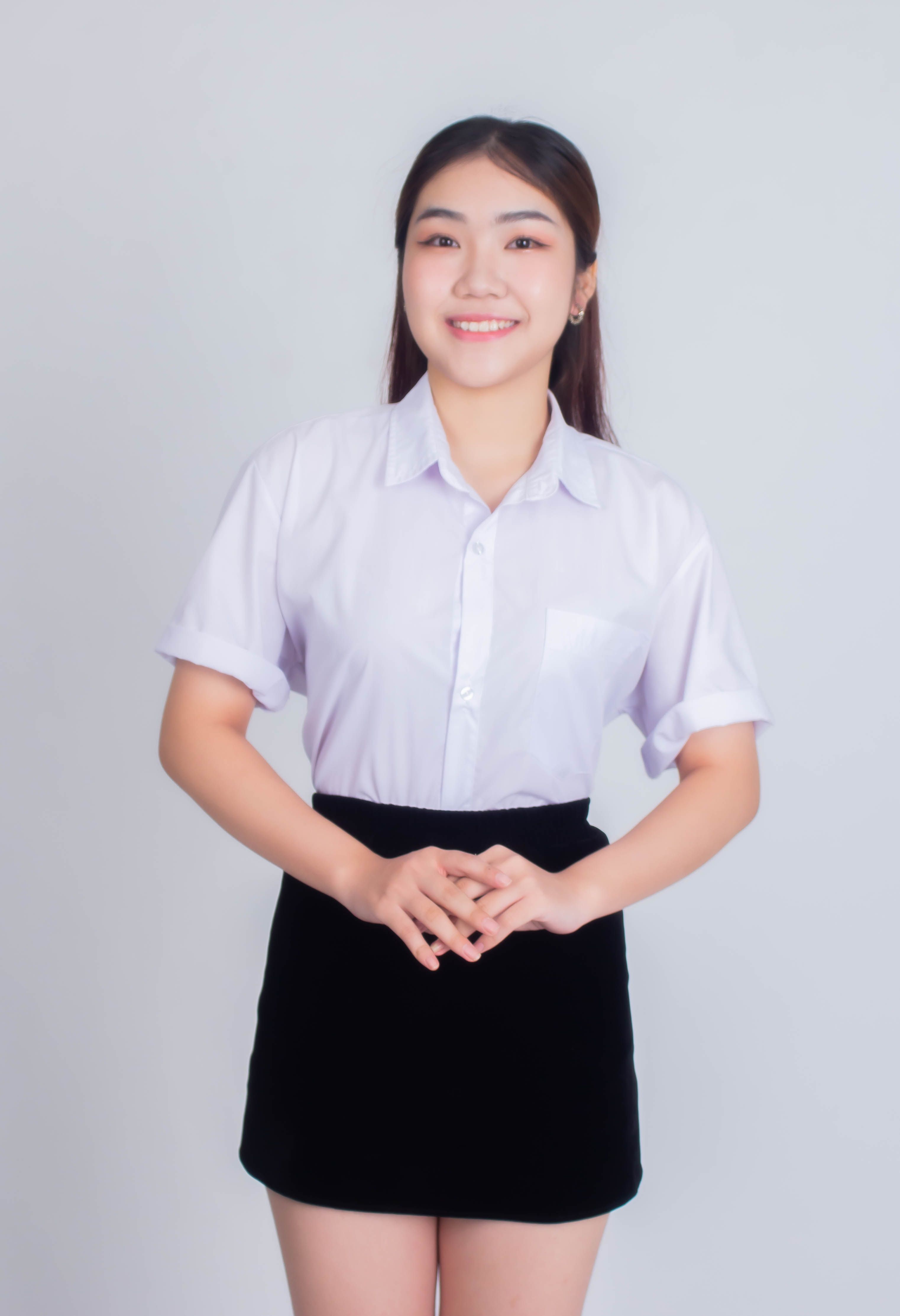 Portrait of a smiling Asian woman standing in a studio wearing a white shirt and black skirt against a white background.