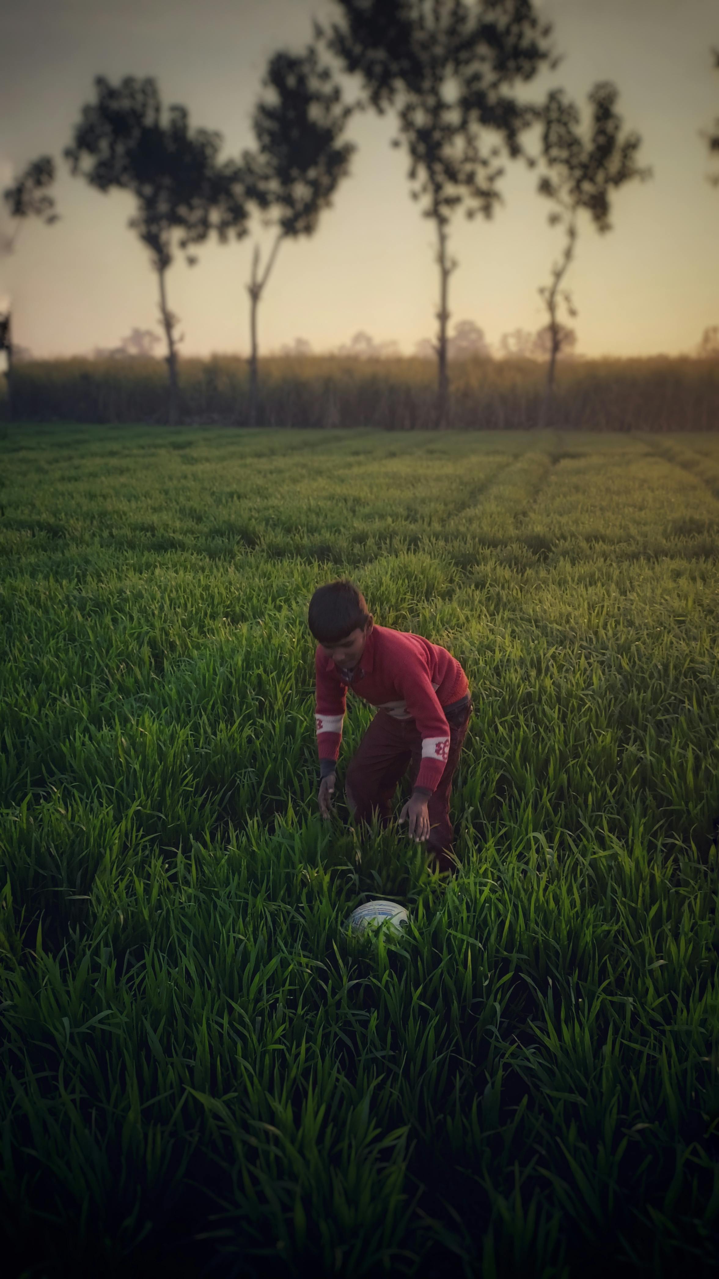 A young boy bends over in a lush green field in India, with a football, during sunset.