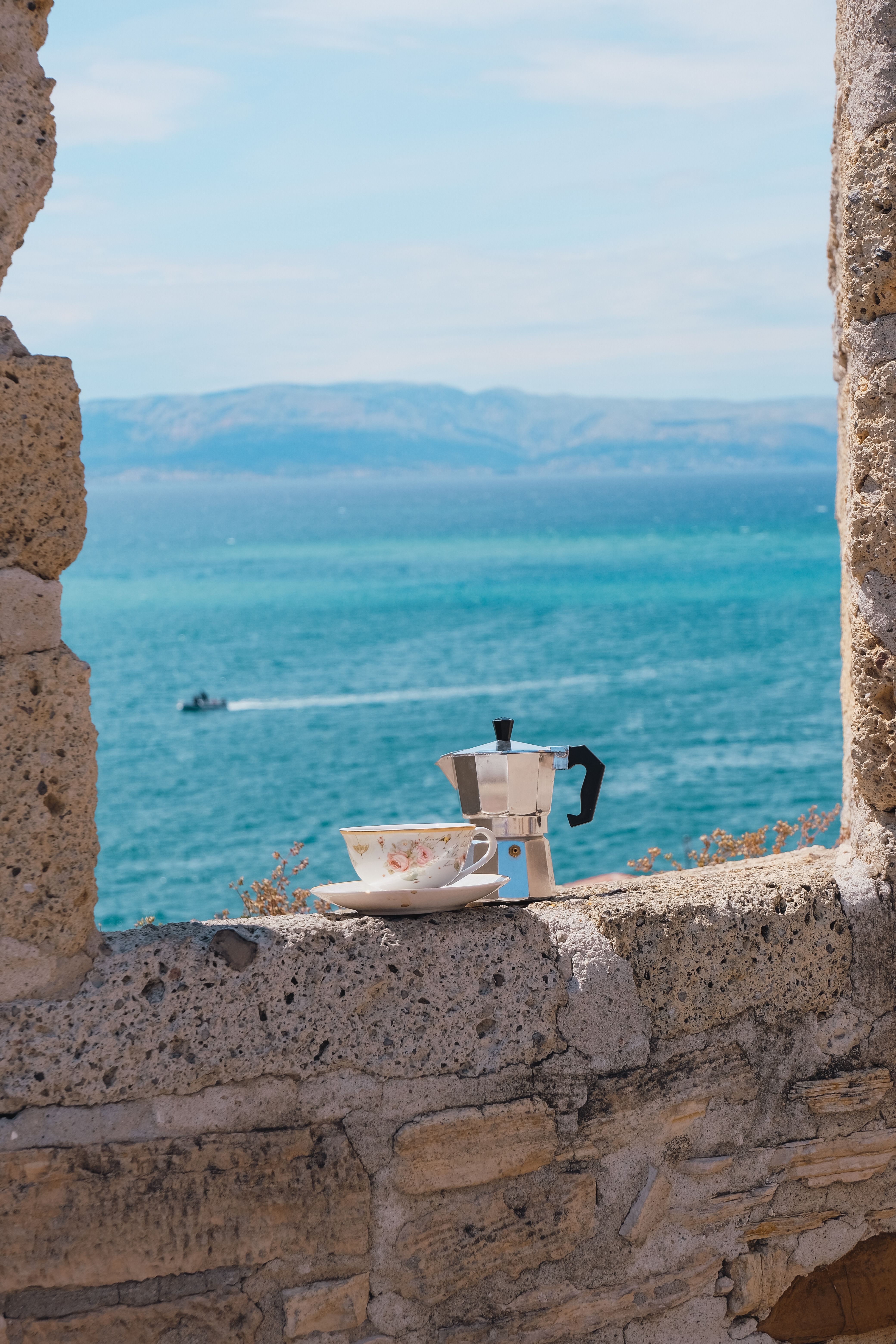 Moka pot and cup sit on a stone wall overlooking the serene blue sea.
