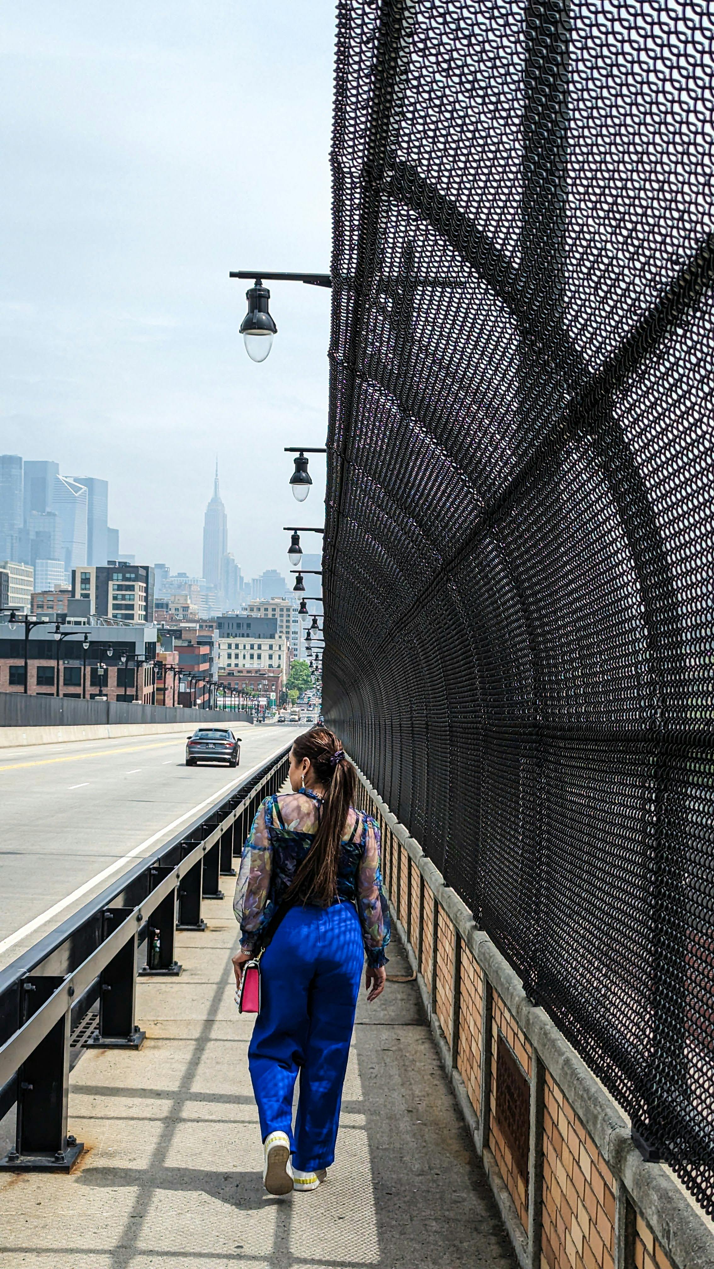 A woman in blue pants walks on a fenced city footbridge with skyline in view.