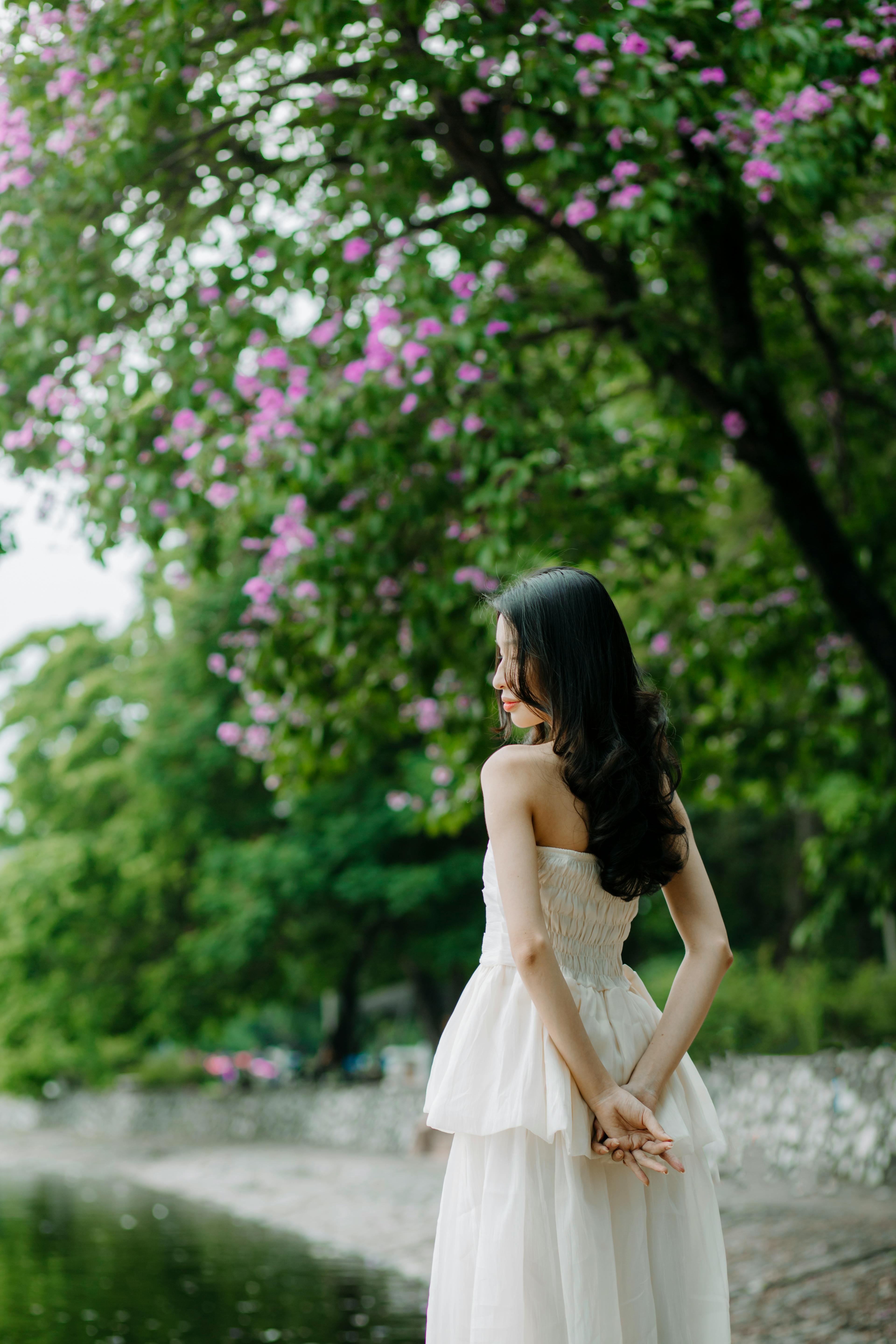 A woman stands gracefully in a white dress amidst vibrant greenery and pink flowers in a park setting.