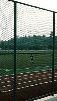 A young child playing on a sports field near a fence, captured from a candid distance.