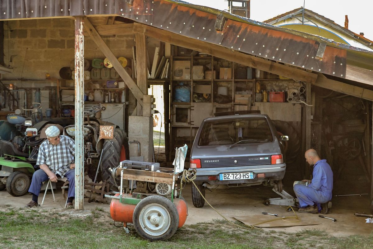 Two men working on a car and old machinery in a rural open-fronted garage