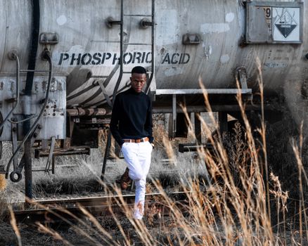 A fashionable man poses by a phosphoric acid tank car on train tracks, showcasing urban style and industrial vibe.