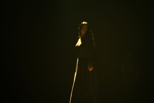 Free A woman in a black dress stands under dramatic lighting in a dark studio setting. Stock Photo