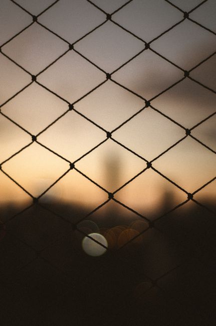 Blurred sunset view through wire fence with bokeh effects, creating an abstract pattern of light and shadows.